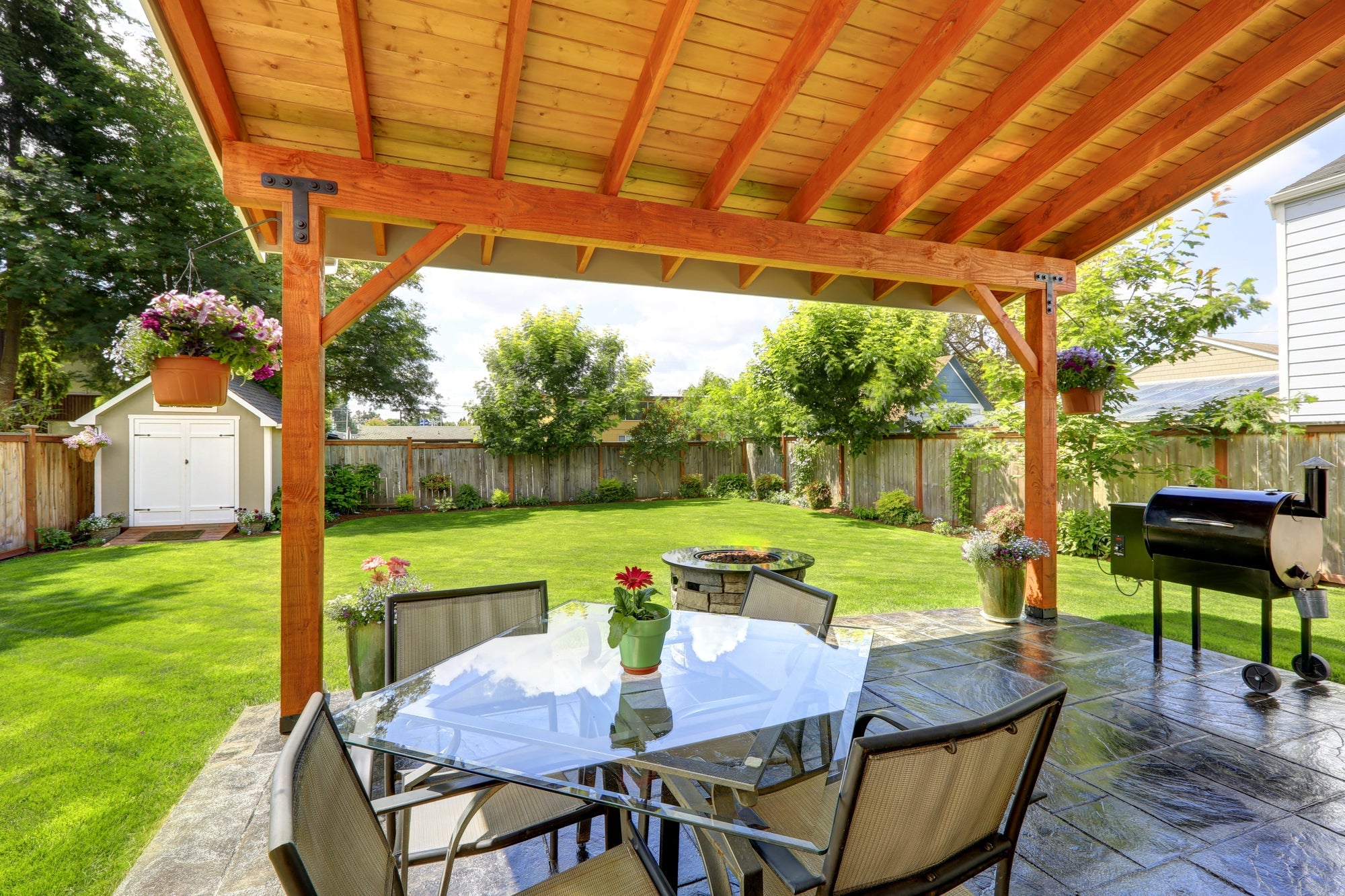 Gartenpavillion selber bauen – Blick unter einem hölzernen Pavillondach auf einen gedeckten Terrassentisch, Grill und eine gepflegte Rasenfläche mit Bäumen und Blumen.